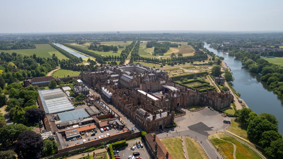 Aerial view of Hampton Court Palace surrounded by lush green gardens and trees, with the River Thames flowing alongside on the right side. The historic brick and stone castle features multiple towers
