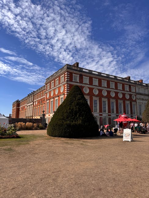 This image shows a large, elegant red brick building with white decorative details and many tall windows, likely part of Hampton Court Palace. In the foreground, there is a neatly trimmed, conical eve