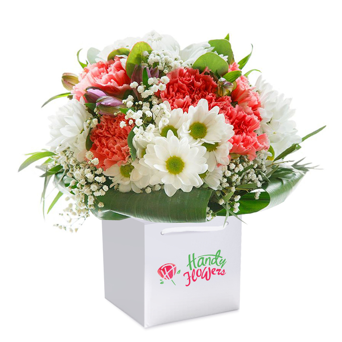 Elegantly poised within a clean, square white gift bag, this radiant bouquet is evocative of the classic beauty cherished in Hampton. The composition begins with lush, ruffled carnations-lovingly selected in shades of coral, peach, and deep red to reflect the dynamic garden palettes seen on walks past Hampton's riverside cottages. Bright white daisies, each centered with a lively yellow-green eye, inject a burst of optimism and contrast, echoing the youthful vibrancy of Bushy Park's open spaces. Amidst these central blooms, wisps of delicate baby's breath infuse cloud-like lightness, while occasional purple-tinged buds peek gently from the ensemble, their presence hinting at hidden nooks and secret gardens dotted across Hampton Hill. Encircling the flowers, broad, glossy green leaves lend strength and form to the arrangement, echoing the canopy of Hampton's ancient trees. The entire floral display is perched within a crisp white bag equipped with sturdy ribbon handles, designed with both practicality and aesthetic refinement in mind-ideal for convenient local gift delivery or pick-up. Rich in texture, colour, and layered detail, this bouquet is thoughtfully crafted by seasoned Hampton florists, capturing both the freshness and celebratory mood suitable for birthdays, anniversaries, or simply to convey heartfelt sentiment to loved ones within the TW12 community and beyond.