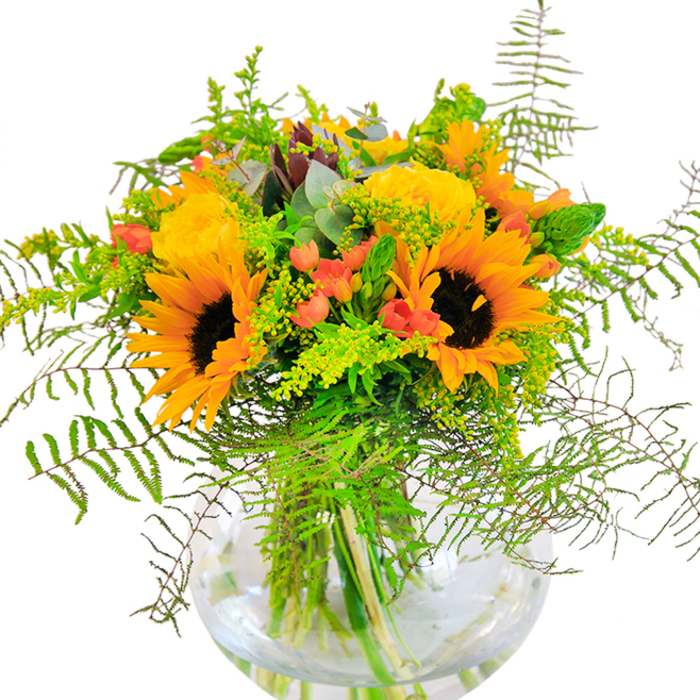This radiant bouquet, created with care by Florist Hampton, is a true celebration of the area's inviting character and natural splendor. At the arrangement's core, brilliant sunflowers bloom, their sun-kissed yellow petals and velvety brown centers evoking memories of picnics and laughter in Hatherop Park. Encircling them, garden-fresh roses unfurl in a buttery yellow, layer upon delicate layer, lending a note of classic romance inspired by the timeless charm of Hampton's riverside cottages. Playful clusters of shiny red Hypericum berries pop against the brighter blooms, conjuring the exuberant palette of local markets in full swing. Luxuriant, flowing fern fronds and golden sprigs of solidago fill out the bouquet, their fine texture and gentle motion reflecting the leafy canopies over Hampton's footpaths. Soft green fillers and slender spikes add structural grace, while silver-blue eucalyptus leaves offer an aromatic touch-cool, clean, and quietly uplifting. Every stem is showcased in a clear, fishbowl-style vase, where they weave together in a fresh, organic tangle grounded in sparkling water. Bathed in gentle daylight, the bouquet radiates a sense of cozy celebration, making it a perfect addition for birthdays, anniversaries, or any occasion to bring Hampton's vibrant spirit into your home.
